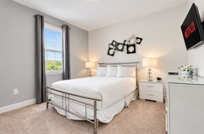 A bedroom with a bed, bedside tables, a desk, a lamp, a wall-mounted TV, and a window with curtains.