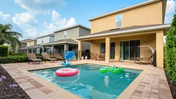Une piscine rectangulaire avec deux bouées gonflables, entourée d’une terrasse en briques et d’un espace extérieur couvert comprenant des chaises et une table.