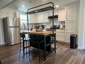 A modern kitchen with white cabinetry, a stainless steel refrigerator, a wooden island with two chairs, and a black trash bin.