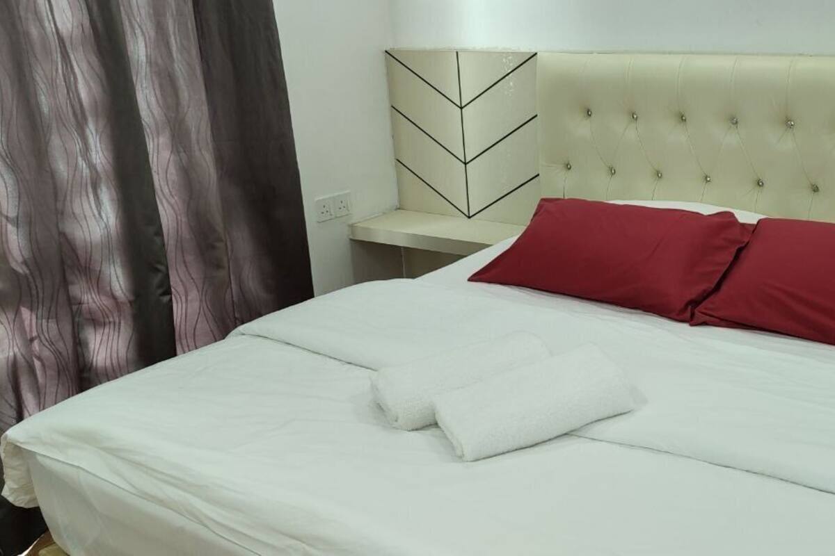 Un lit bien fait, avec du linge de lit blanc et deux oreillers rouges, une tête de lit capitonnée de couleur crème et un motif géométrique moderne sur le mur.