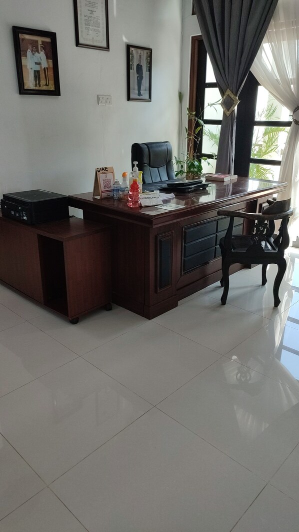 A spacious office with a large desk, a chair, and a potted plant. There are framed pictures on the wall and a window with curtains.