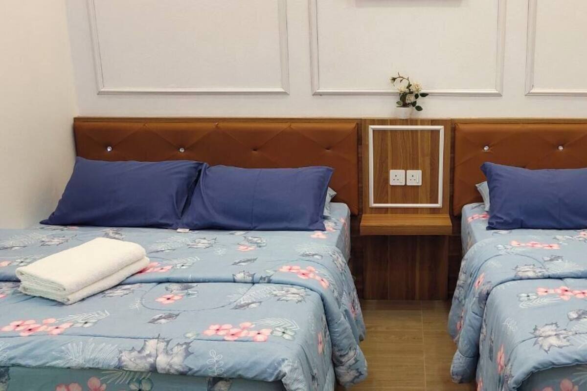 Une chambre d’hôtel avec deux lits, chacun agrémenté de draps à motifs floraux et d’oreillers bleus. Chaque lit est doté d’une tête de lit en bois et d’une petite table de chevet en bois, avec une prise électrique située entre les deux lits.