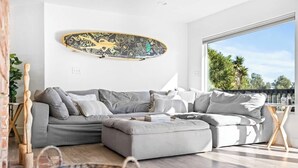 A modern living room with a grey sectional sofa, a skateboard on the wall, a wooden side table, and a view of palm trees through a large window.