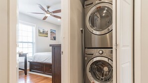 A laundry room with two stacked front-loading washers and dryers, a ceiling fan, a bed with a wooden headboard, and a window with blinds. - Central Convenient Home | Free Parking | Near U St (Washington)