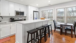 A modern kitchen with white cabinetry, a central island with bar stools, a dining area with a table and chairs, and large windows with blinds. - Central Convenient Home | Free Parking | Near U St (Washington)