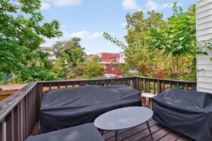 A covered outdoor seating area with a table and chairs, surrounded by a wooden deck and foliage.