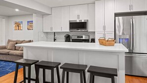 A modern kitchen with white cabinets, a stainless steel refrigerator, a microwave, and a coffee maker. There are three bar stools in front of the kitchen island. - Central Convenient Home | Free Parking | Near U St (Washington)