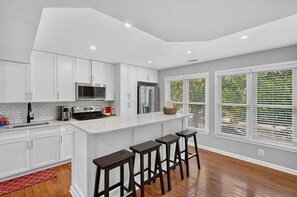 A modern kitchen with white cabinets, a central island with stools, and a view of greenery outside. - Central Convenient Home | Free Parking | Near U St (Washington)