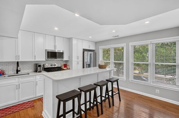 A modern kitchen with white cabinets, a central island with stools, and a view of greenery outside.