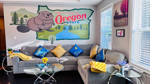 Una sala de estar con sofá, almohadones, una mesa de centro de vidrio y un mural que da la bienvenida a los visitantes a Oregon State.