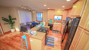 Una cocina moderna con armarios de madera, estufa a gas y refrigerador. Hay un comedor con una mesa y sillas, y una planta en maceta.