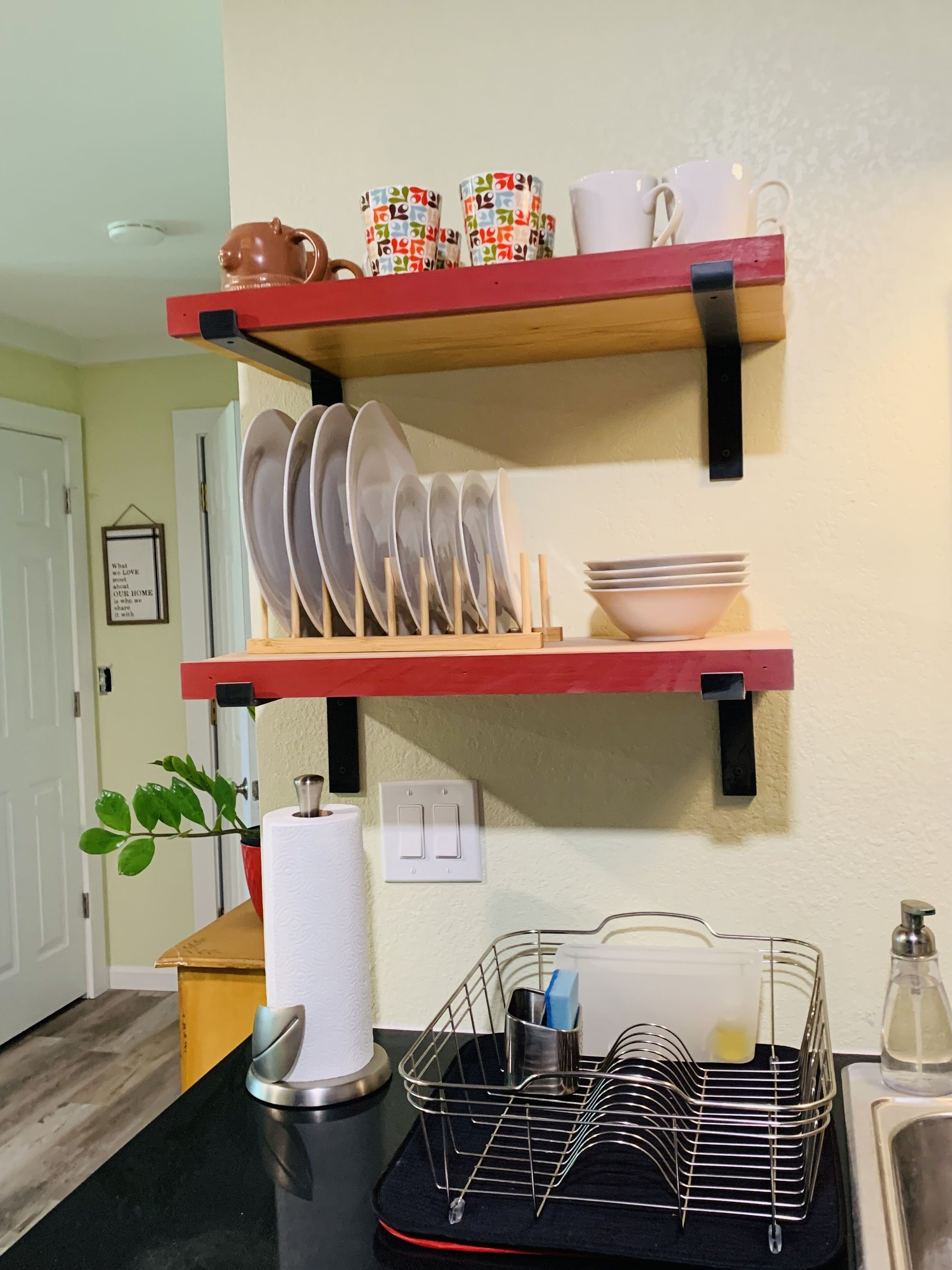 A kitchen with two wooden shelves holding plates, bowls, and mugs. There is a black tray with utensils and a plant on the counter.