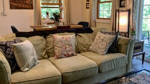 Ein gemütliches Wohnzimmer mit einem beigefarbenen Sofa, einem Esstisch mit Stühlen und einem großen Fenster mit Vorhängen.