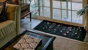 Ein Zimmer mit einer großen Glasschiebetür, die zu einem überdachten Außenbereich führt, ein Sofa, ein kleiner Tisch mit einem dekorativen Gegenstand und ein Teppich mit hängenden Verzierungen.