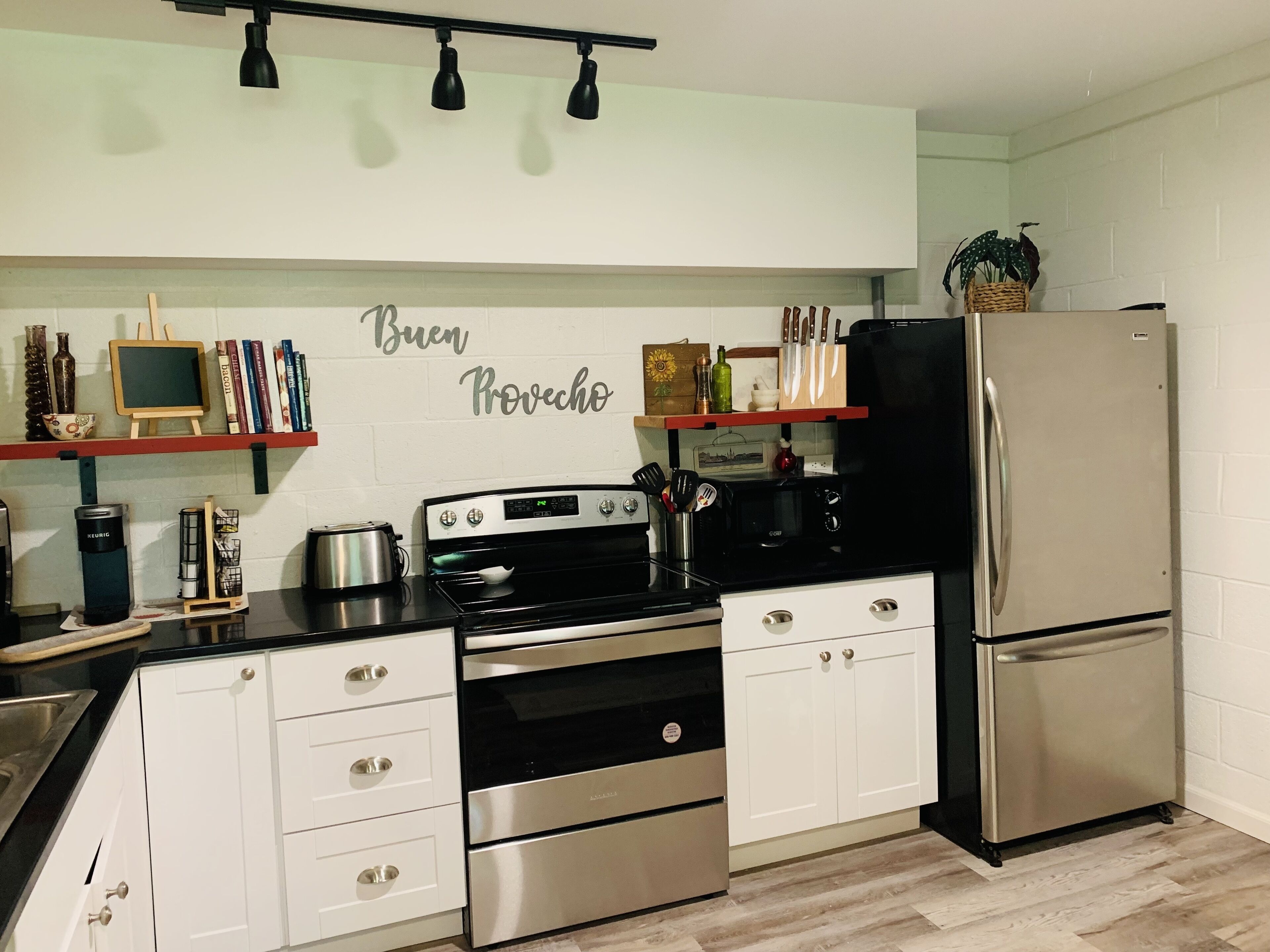 A kitchen with white cabinets, a stainless steel refrigerator, a black stove, and a wall-mounted shelf with books and kitchen utensils.