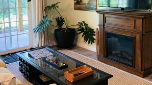 Ein Wohnzimmer mit Kamin, Fernseher, einer Uhr und einem großen Fenster mit Vorhängen.
