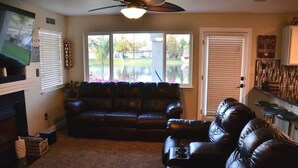 A living room with a black leather sofa, a ceiling fan, a fireplace, and a large window with a view of a lake.