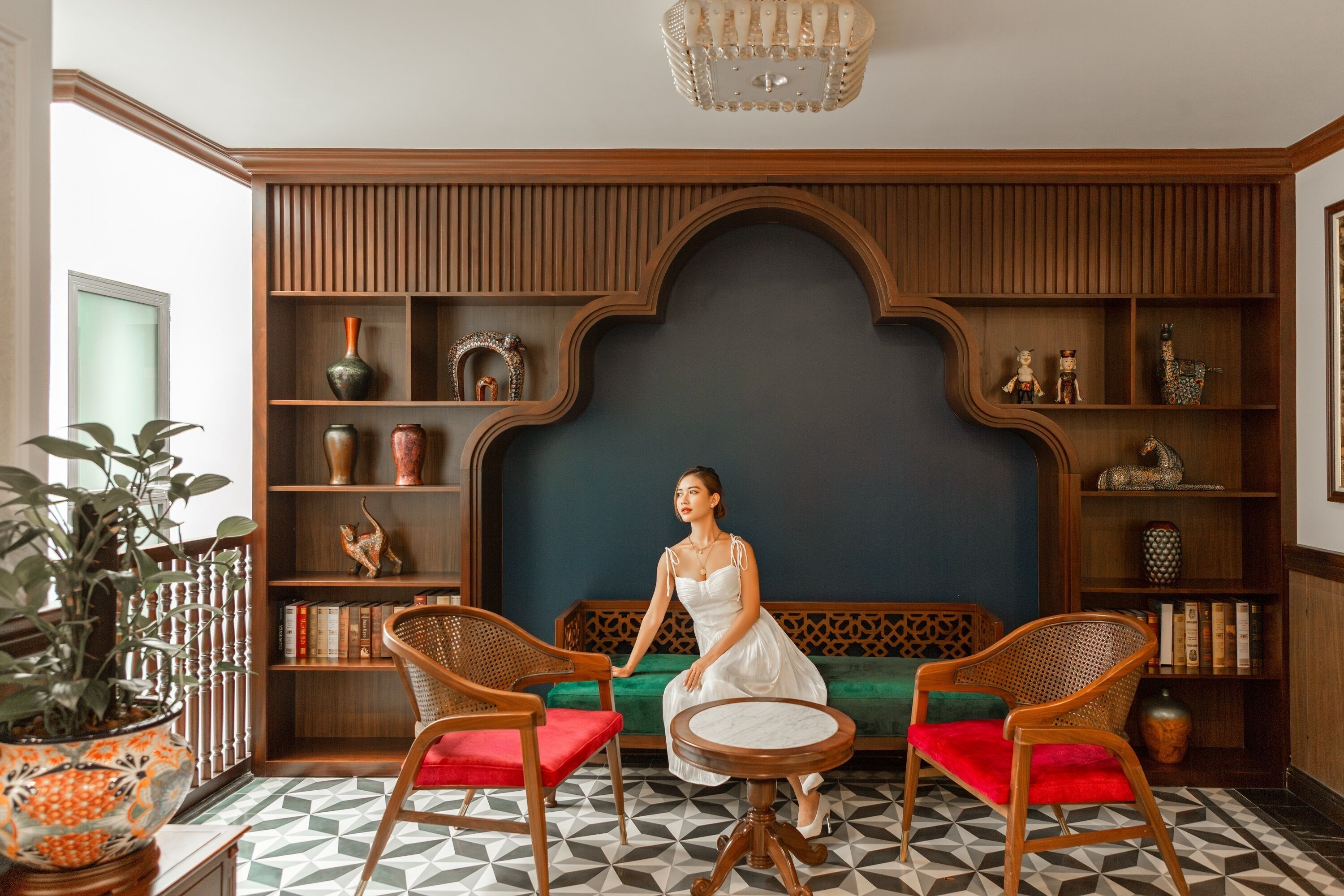 A woman sitting on a green sofa in a room with wooden shelves, a patterned floor, and a chandelier.