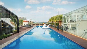 A swimming pool with a glass enclosure on one side, a modern building with a curved facade on the other, and a clear sky with clouds. - Grand Fleuve Boutique  (Bai Chay)