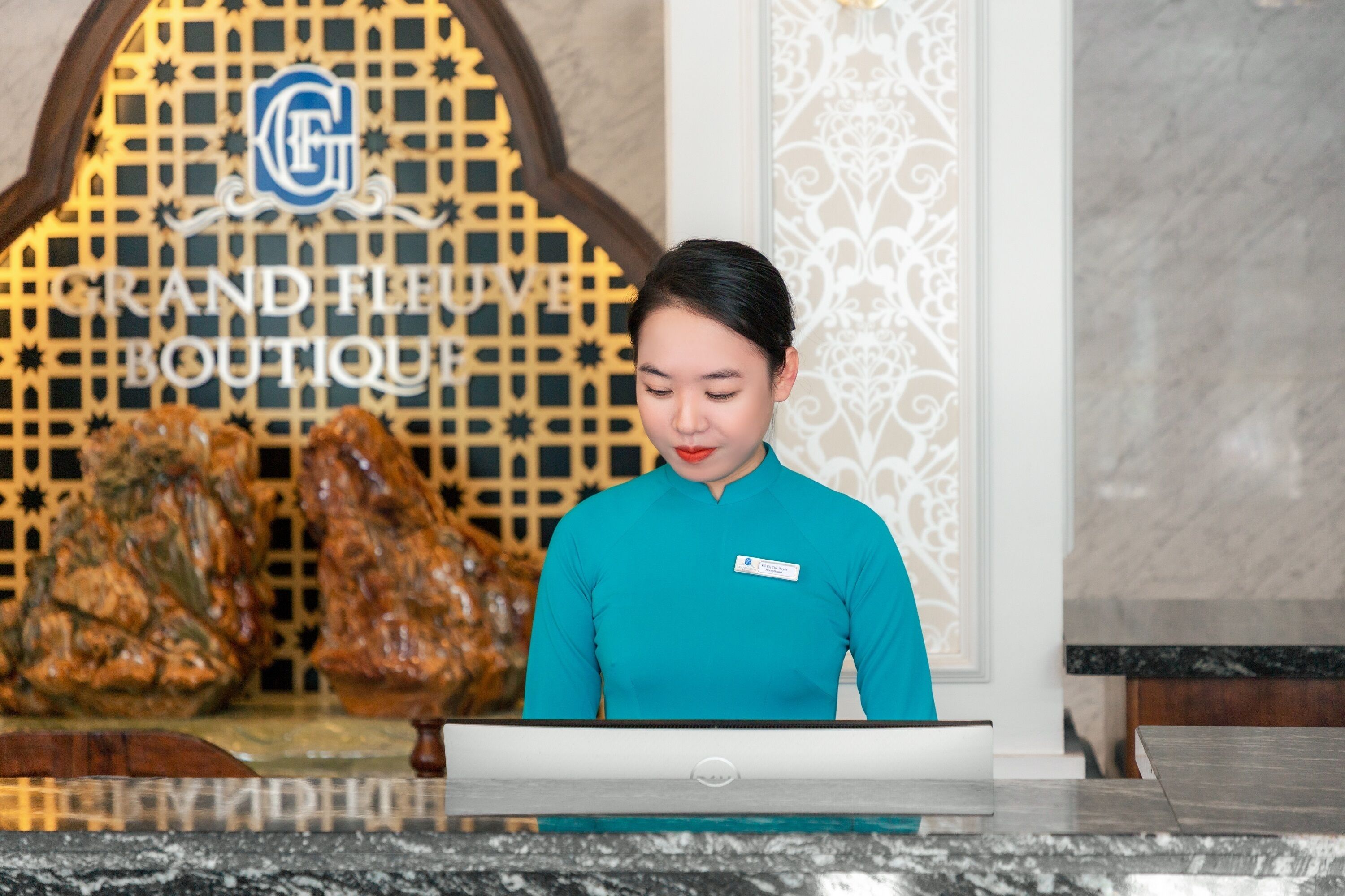 En receptionist vid hotellreceptionen, med en skylt i bakgrunden som visar texten ”GRAND FLUVE BOUTIQUE”.