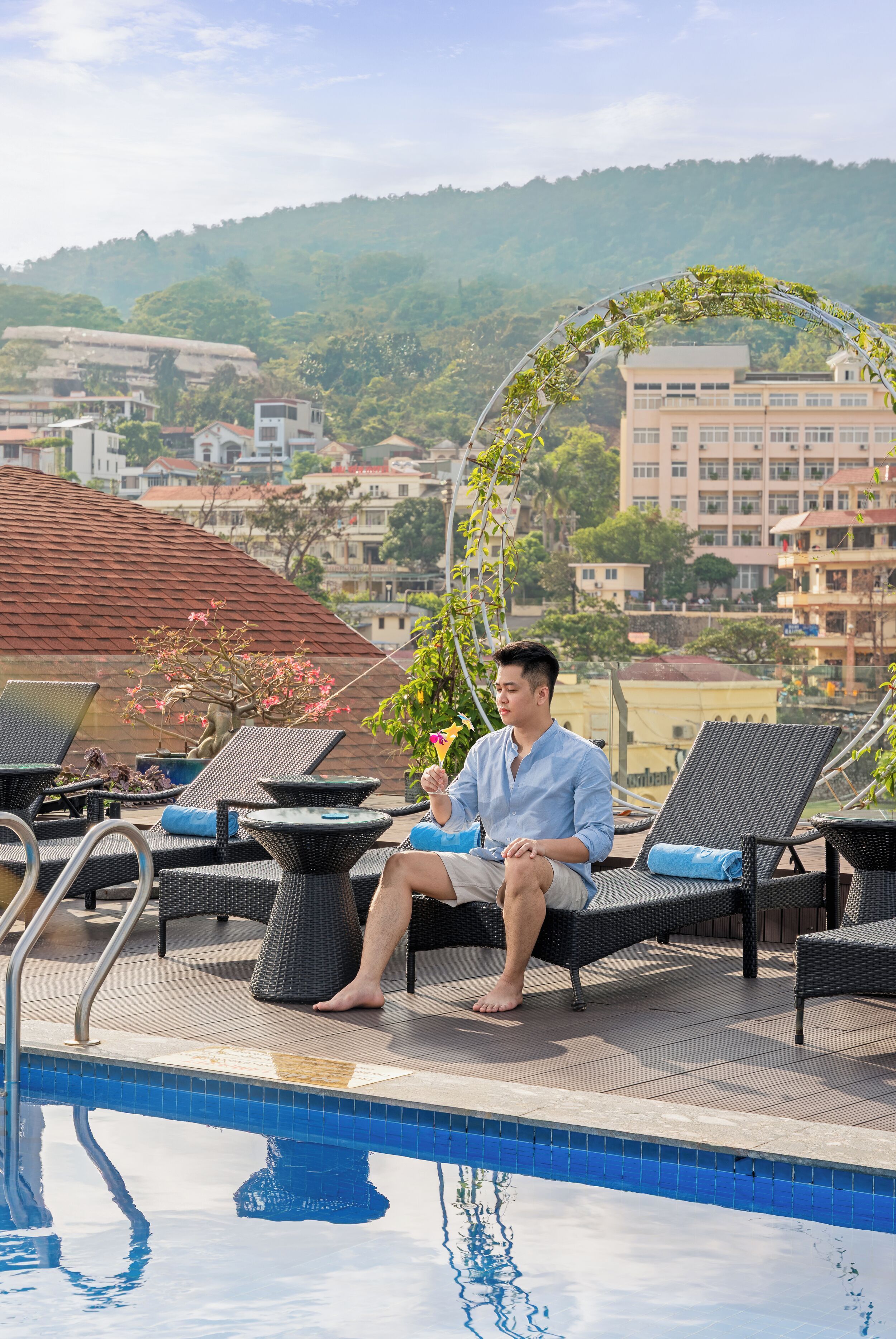 A man sitting by a pool with lounge chairs and a decorative archway.
