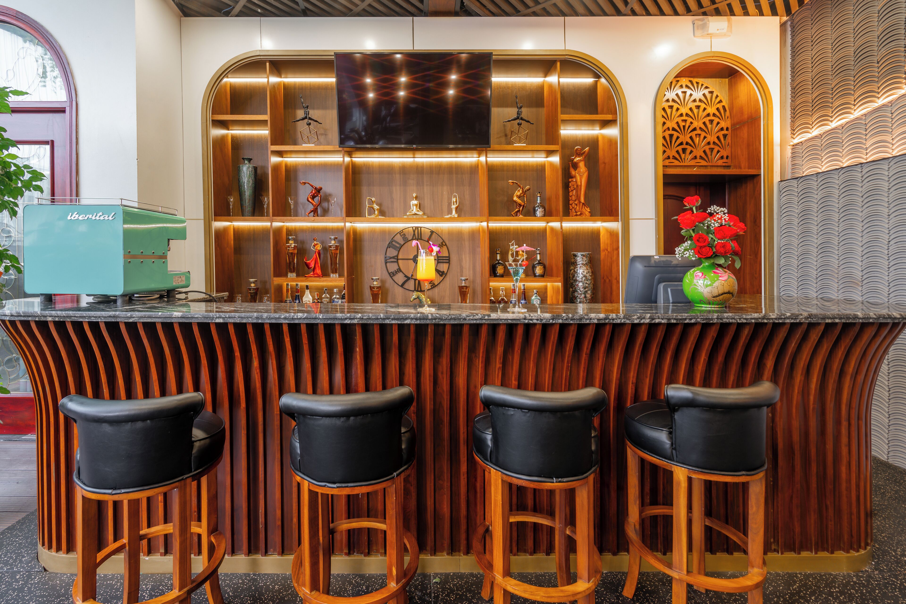 Un espace bar avec un comptoir en bois, trois tabourets de bar noirs, une horloge et des étagères contenant diverses bouteilles et objets décoratifs.
