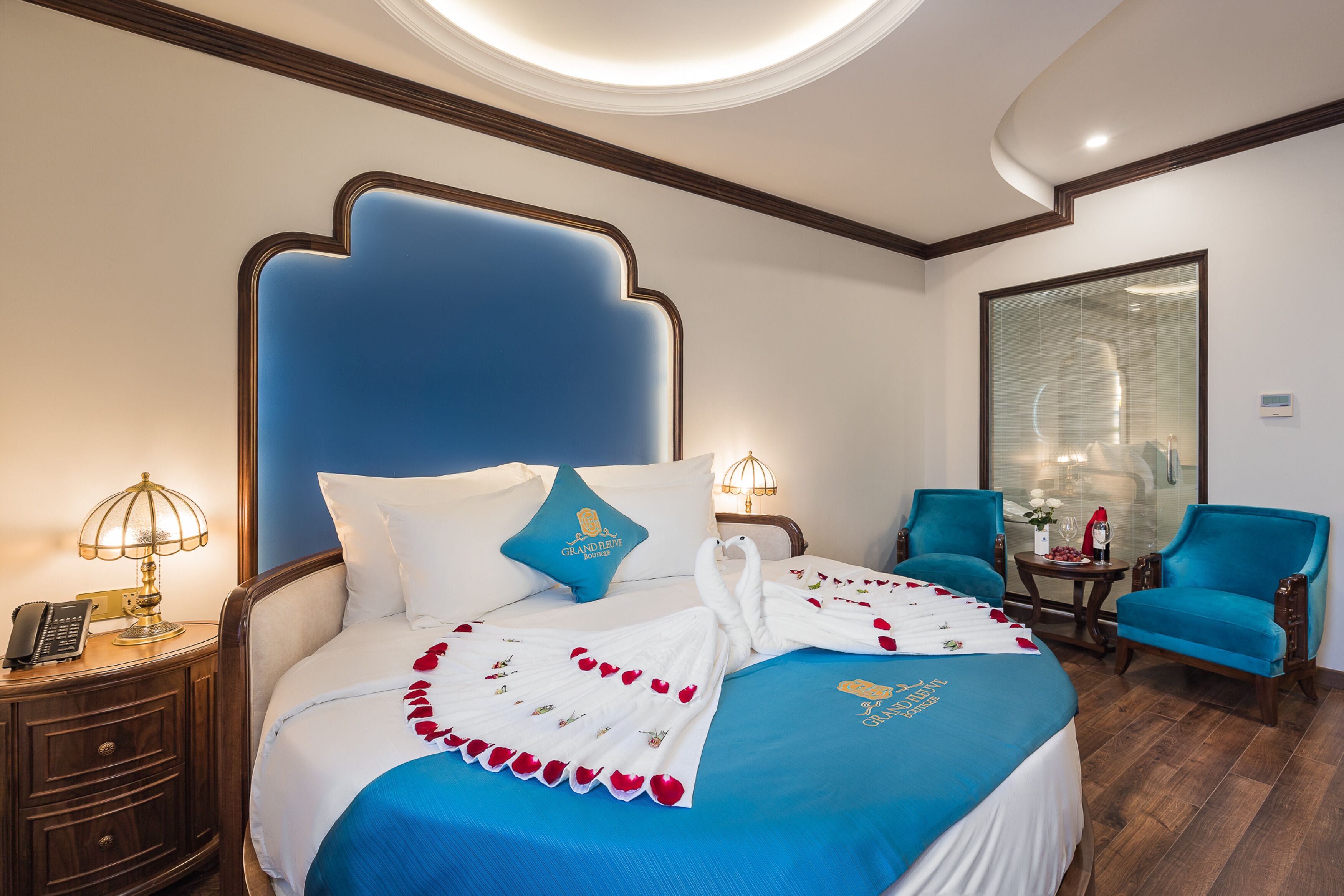 A hotel room with a large bed, a nightstand with a lamp, a small table with a vase, and a blue armchair.