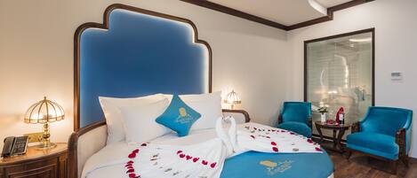 A hotel room with a large bed, a nightstand with a lamp, a small table with a vase, and a blue armchair.