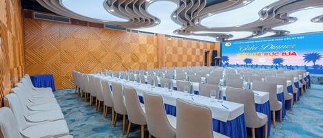 A conference room with a long table set for a meeting, a stage with a presentation screen, and a patterned ceiling.