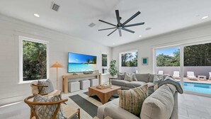 A spacious living room with a large flat-screen TV, a ceiling fan, and a pool visible through the glass doors. - Barton Vista Del Mar | Paradise Perfected!! | Deeded Access with Free Beach Chairs & Umbrellas 100 Yards Away! (Miramar Beach)