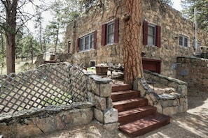 Traditional stone building with a wooden staircase and metal fence, set amidst lush vegetation.