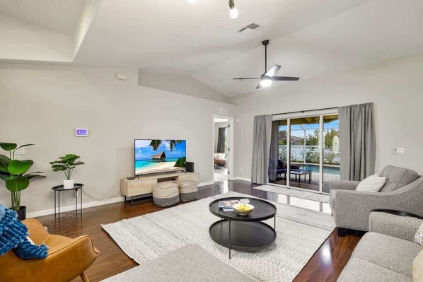 A modern living room with a flat-screen TV, a grey sofa, a coffee table, and a view of the outdoors through large sliding glass doors.