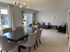 Et spiseområde med et mørkt træbord og seks stole, en grå sofa med farverige puder, en sort lænestol og en moderne lysekrone.