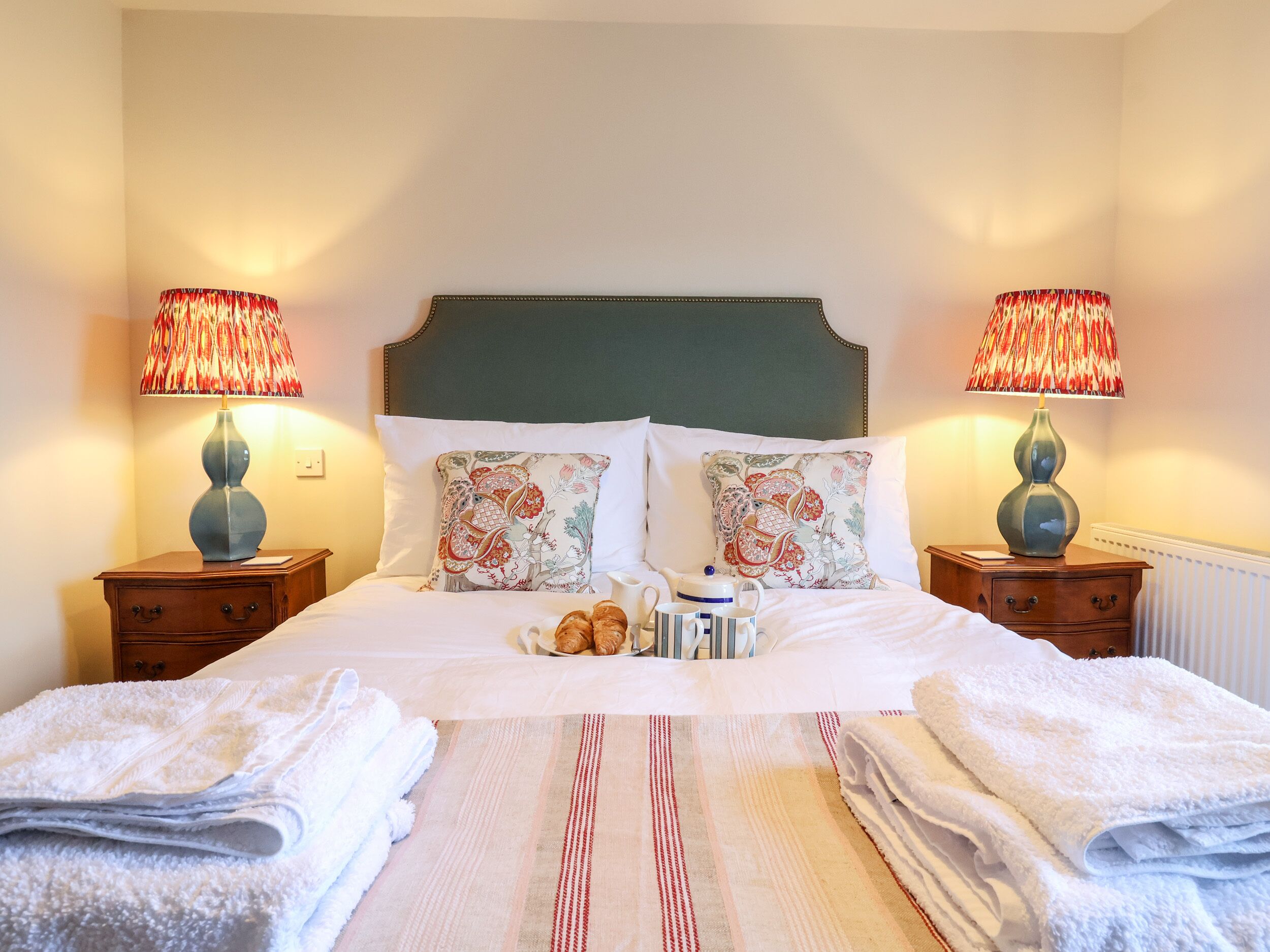 A neatly made bed with white linens, two decorative lamps, and a bedside table with a vase.