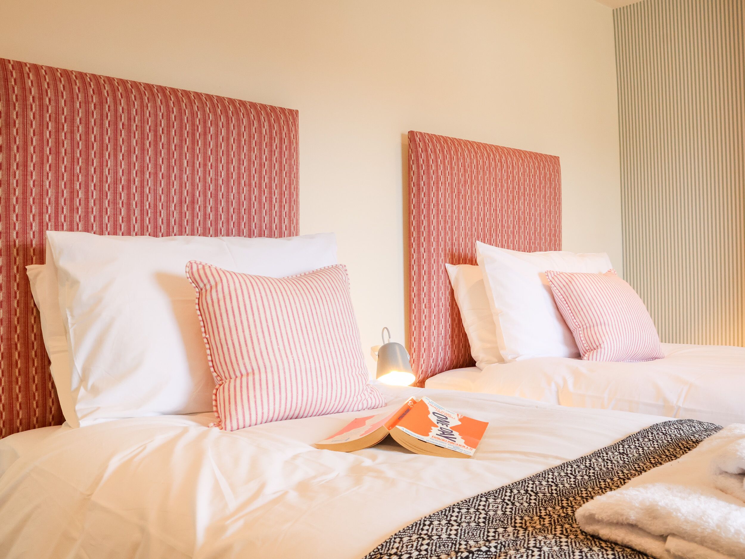 A hotel room with two beds, each with white bedding and striped pillows. There is a book on the bed and a patterned throw blanket.
