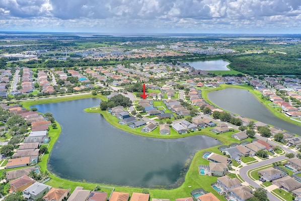 An aerial view of a residential area with multiple houses, a large central lake, and a red arrow pointing to a specific location. - Gilded Parasol By Shine Villas Remington Golf #410 4 Bedroom Home (Kissimmee)