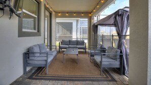 A covered patio with a gray sofa set, a small table, and string lights. - Marvel Mansion! Championsgate Resort Shine Villas 9 Bedroom Villa (Davenport)