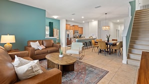 A spacious living room with a brown leather sofa, a coffee table, and a dining area with chairs and a table. A kitchen is visible in the background. - Lazy Life At Storey Lake By Shine Villas #901 5 Bedroom Condo (Kissimmee)