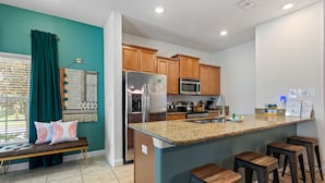 A modern kitchen with wooden cabinets, a stainless steel refrigerator, a microwave, and a granite countertop. There is a bench with cushions and a decorative wall hanging. - Lazy Life At Storey Lake By Shine Villas #901 5 Bedroom Condo (Kissimmee)