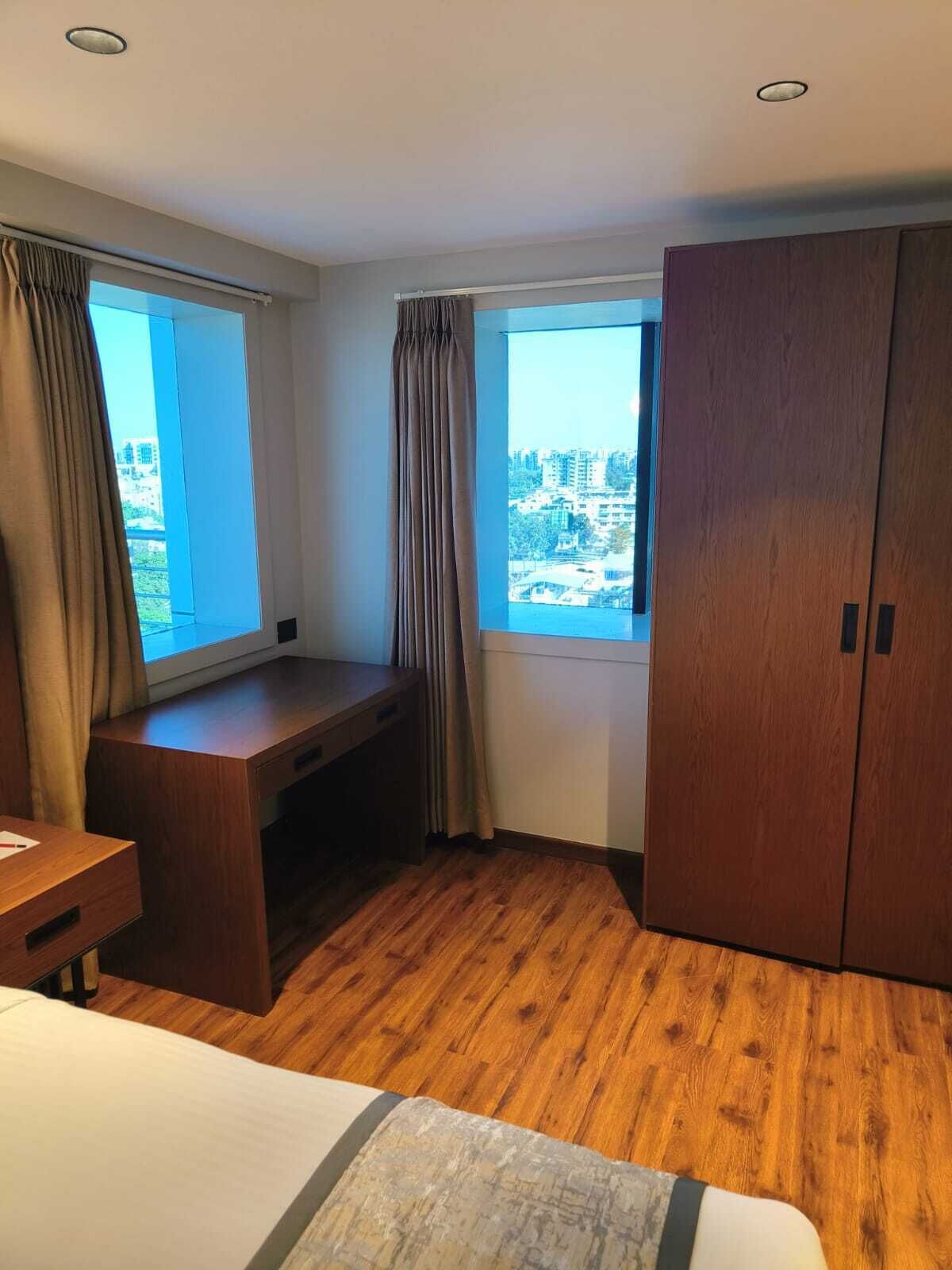 Une chambre d’hôtel avec un bureau en bois, deux fenêtres avec des rideaux, une armoire et un lit.