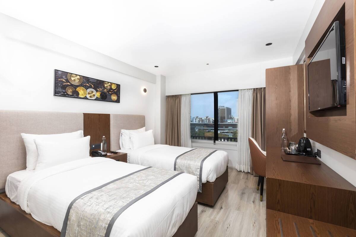 Habitación de hotel con dos camas, televisor, escritorio y un ventanal con vistas a la ciudad.