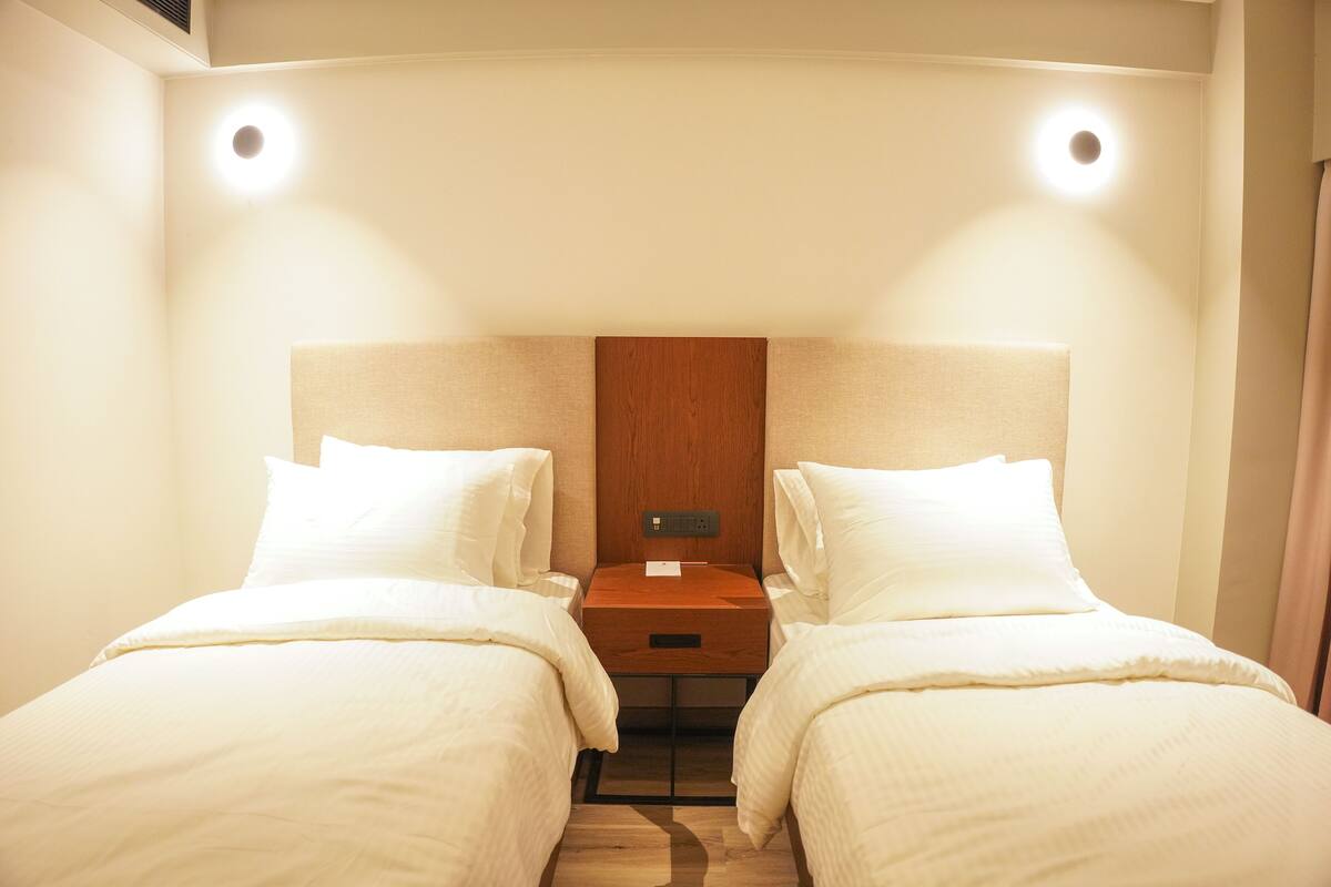 Habitación de hotel con dos camas, cabecera de madera, mesita de noche e iluminación en la pared.