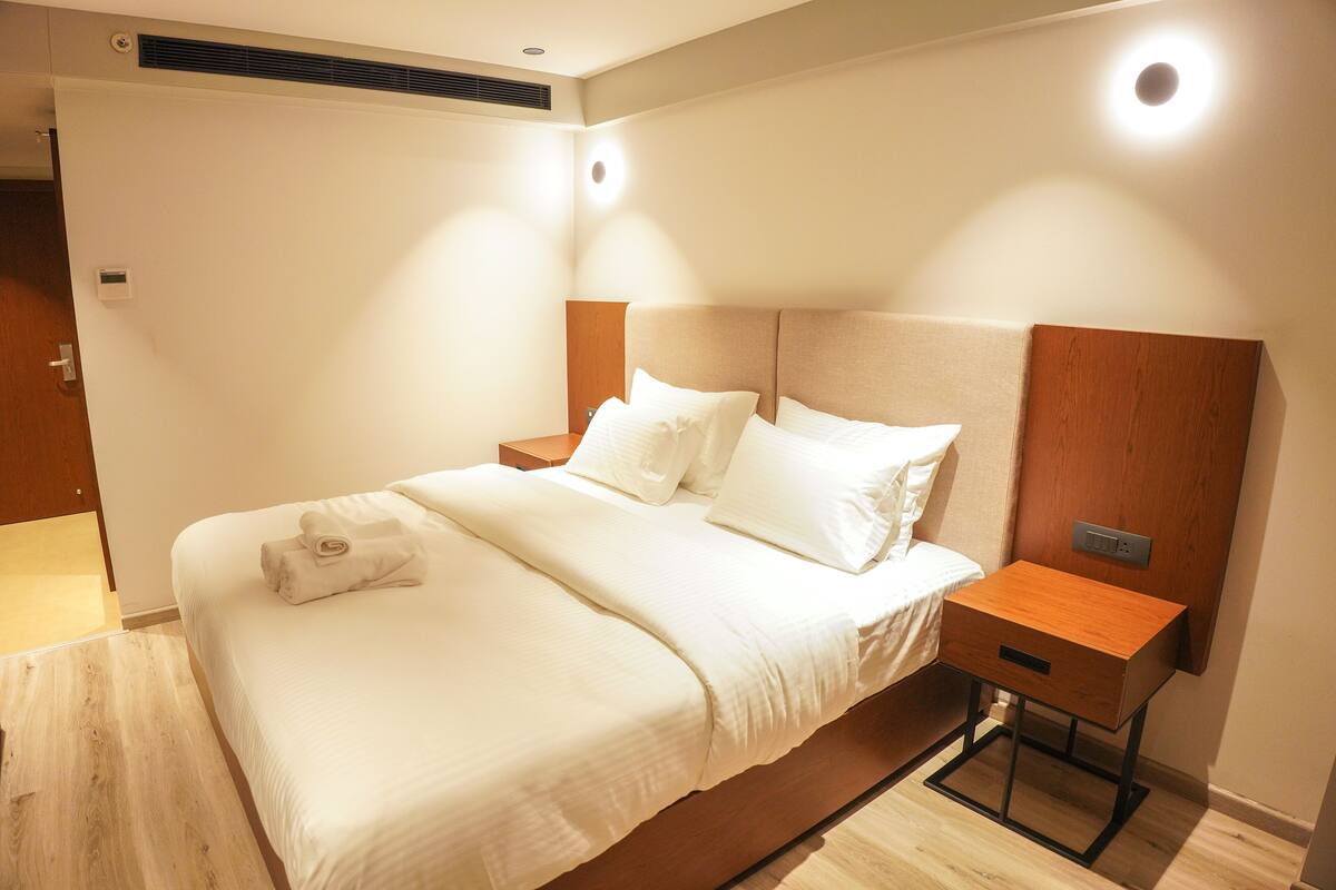 Una cama bien hecha con sábanas blancas, una mesita de noche de madera y una lámpara de pared.