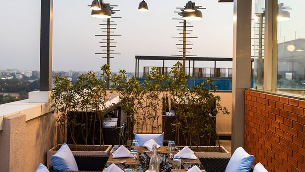 A rooftop dining area with wicker furniture, white cushions, and a long table set for a meal. - Ramee International Surat (Surat)