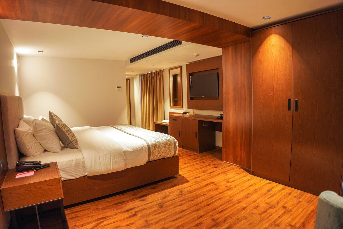 Una habitación de hotel moderna con piso de madera, una cama grande con ropa blanca, un escritorio con silla, un televisor montado en la pared y un armario.