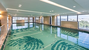 A large indoor swimming pool with clear water, surrounded by walls with large windows offering views of the city. - Ramee International Surat (Surat)