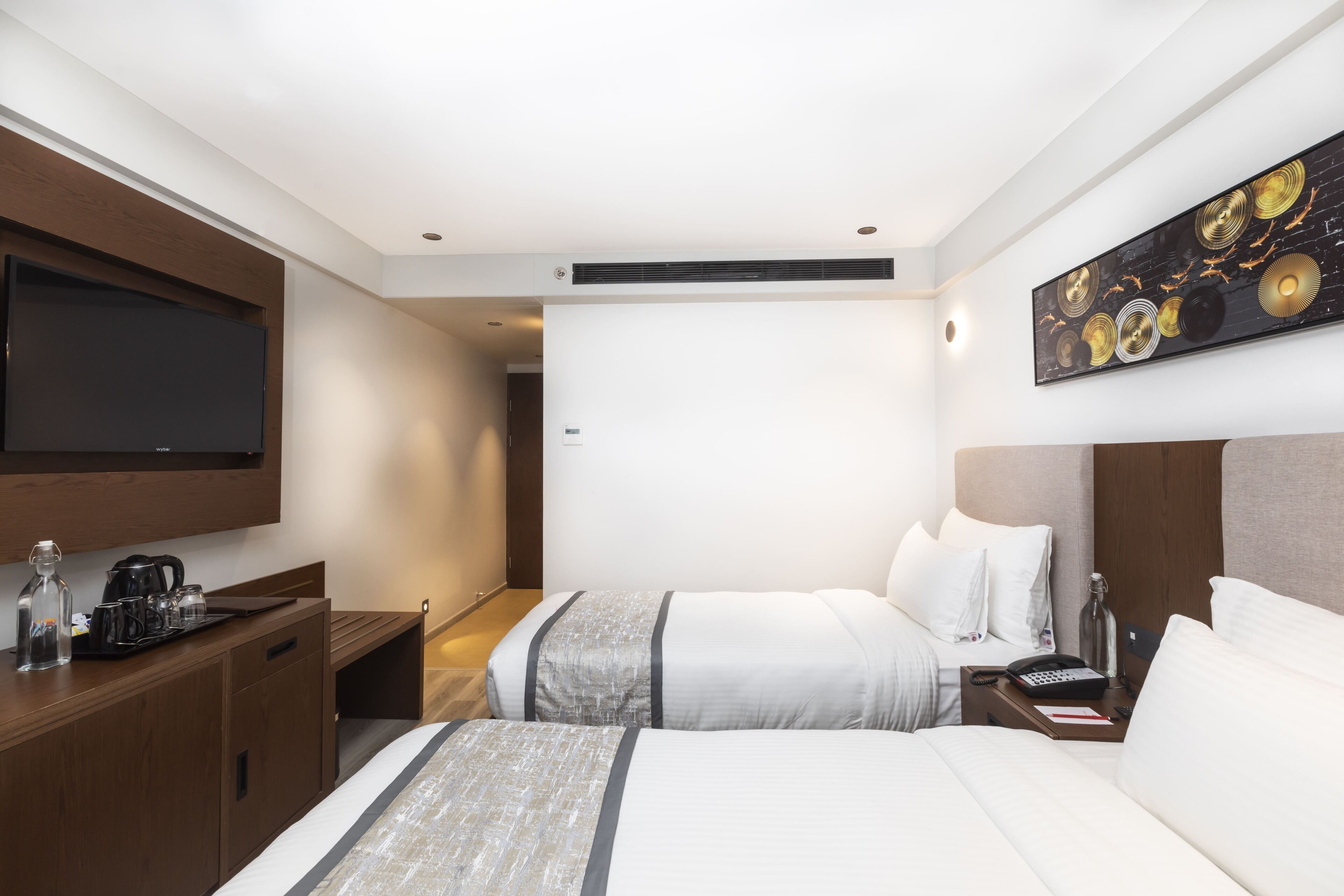 A hotel room with a flat-screen TV mounted on the wall, a wooden desk with a coffee maker, two beds with white linens, a painting on the wall, and a nightstand with a lamp and phone.