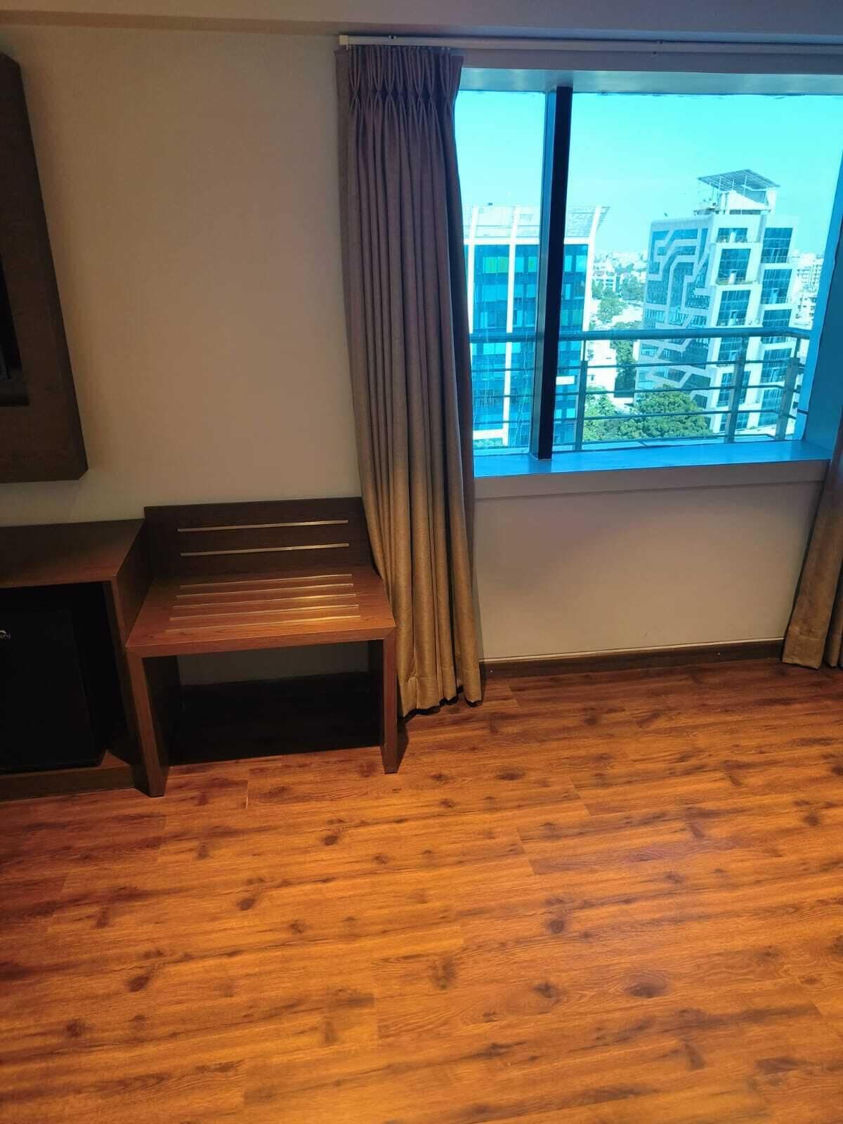 Une pièce dotée d’une grande fenêtre, d’un plancher en bois, d’un banc et offrant une vue sur des immeubles modernes.