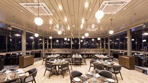 A spacious restaurant with multiple dining tables, high ceilings, and large windows offering views of the cityscape at night. - Ramee International Surat (Surat)
