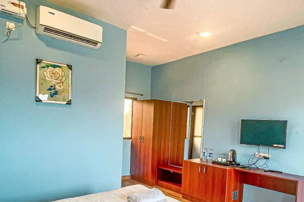 A bedroom with a bed, a ceiling fan, an air conditioning unit, a wooden wardrobe, a vanity with a mirror, and a television on the wall.